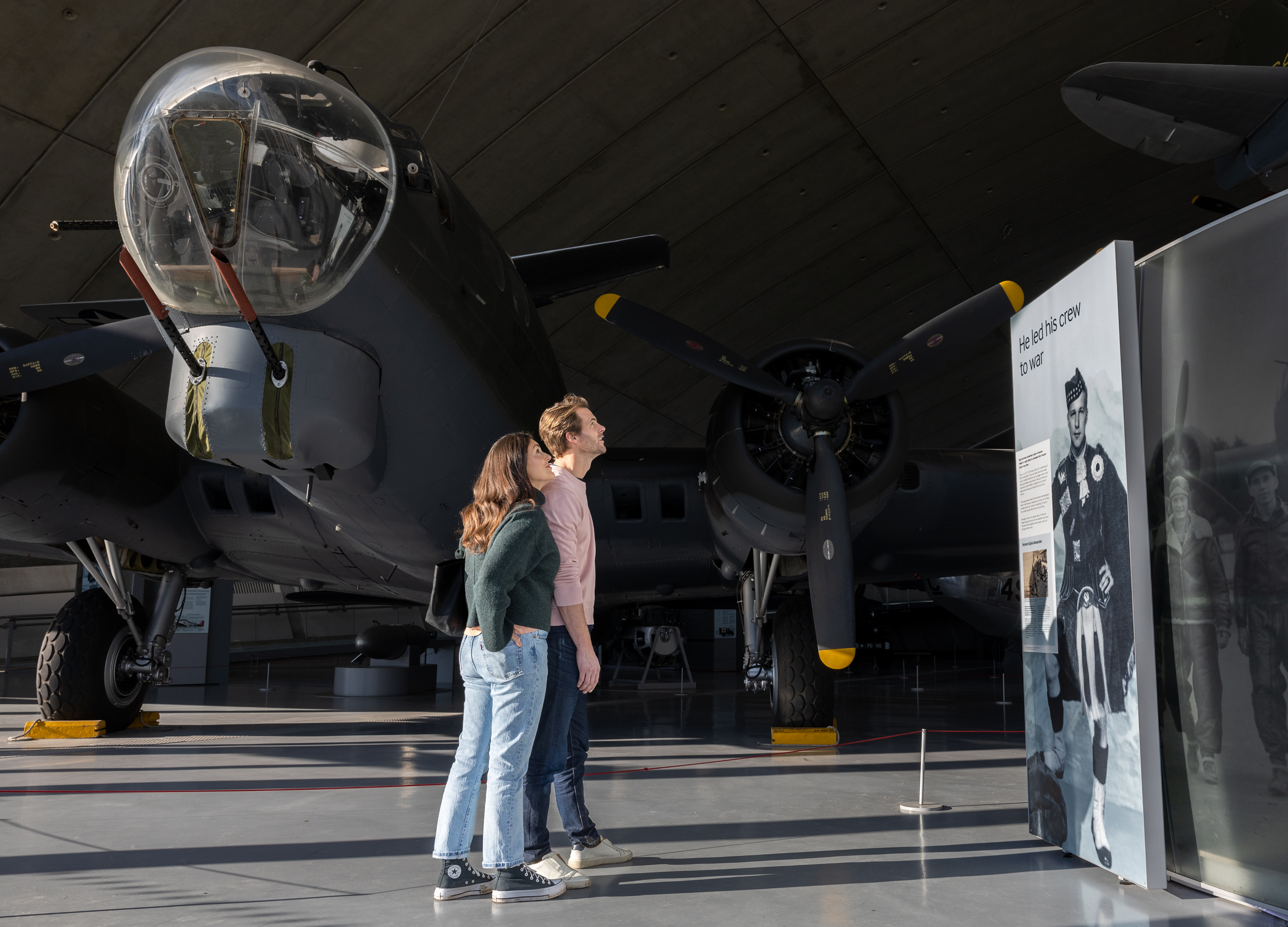 A young couple looking at a display in the American Air Museum with the B-17 Flying Fortress in the background